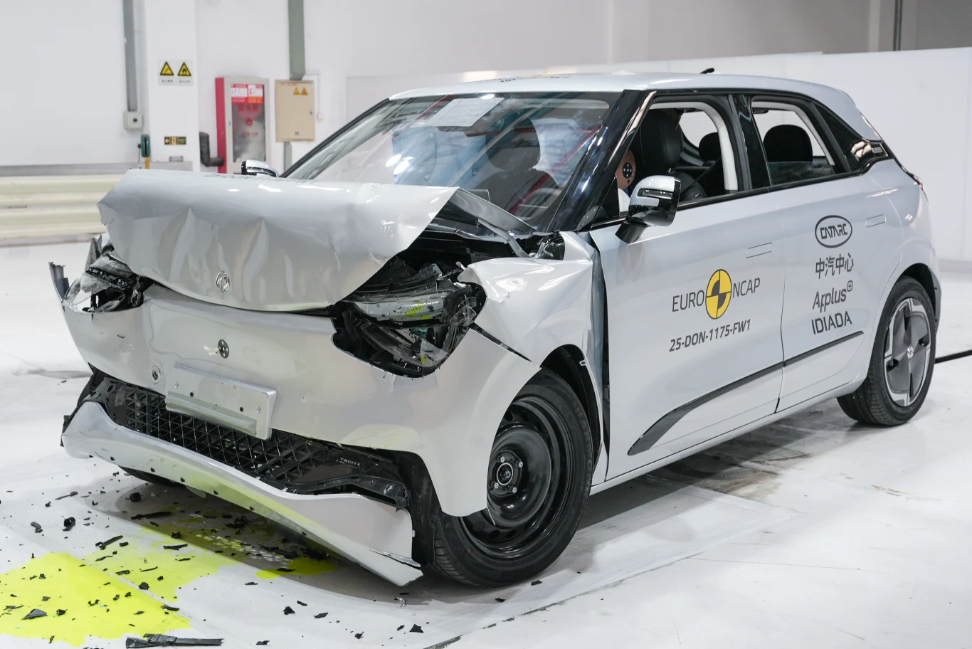 Samochód osobowy o białym kolorze z silnie uszkodzonym przodem, stojący w pomieszczeniu testowym. Na karoserii widoczne oznaczenia instytucji Euro NCAP oraz napisy po chińsku, informujące o przeprowadzanym teście zderzeniowym.