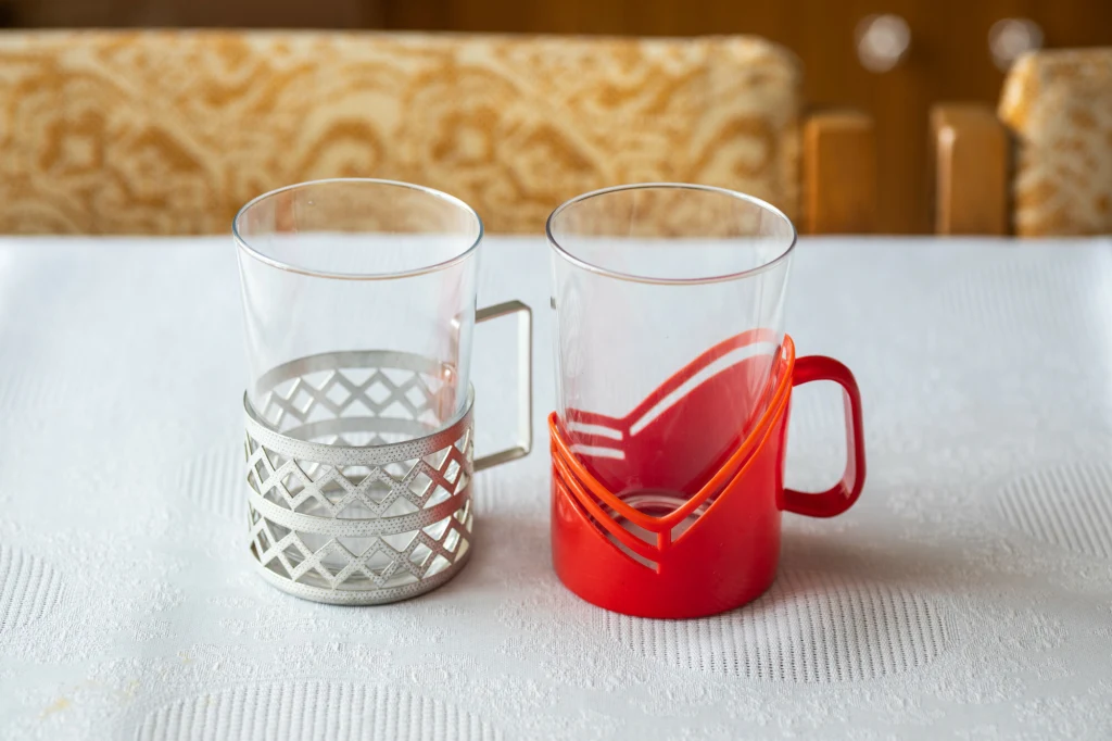 Szklanka tzw. koszyczku cieszyła się ogromną popularnością w czasach PRL-u Dwie szklanki z uchwytami stojące na białym obrusie, jedna w metalowym koszyczku, druga w czerwonym plastikowym uchwycie.