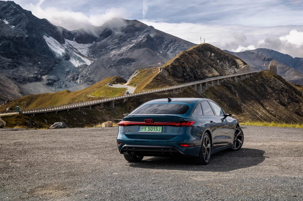 Nowoczesny samochód Audi o ciemnoniebieskim kolorze zaparkowany na tle malowniczych gór, krętej drogi i mostu, z dynamiczną linią horyzontu oraz częściowo zachmurzonym niebem.