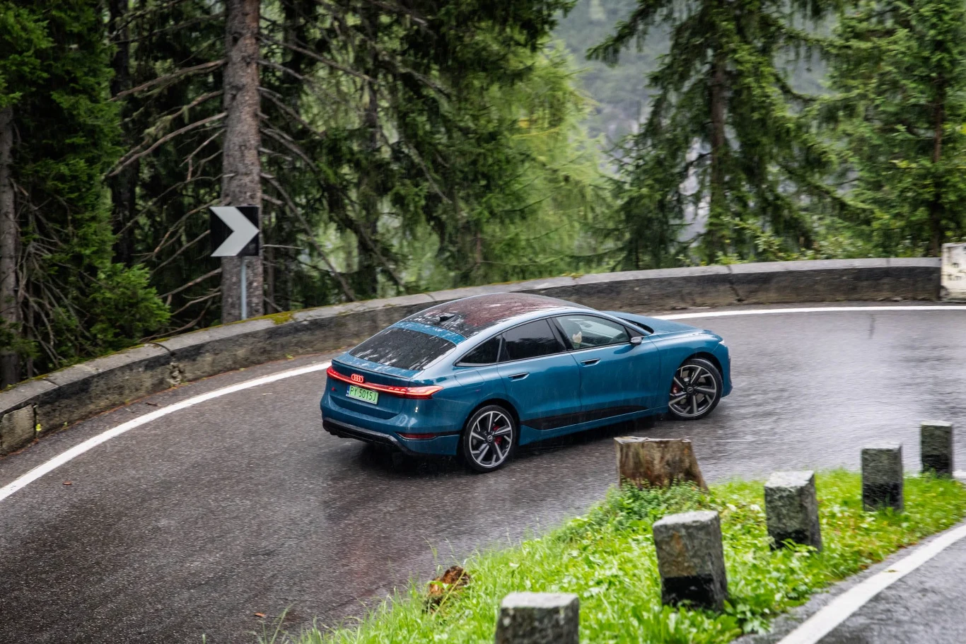 Na mokrym asfalcie Tyrolu pneumatyczne zawieszenie i napęd quattro pokazują pełnię możliwości Nowoczesny niebieski samochód pokonuje ostry, mokry zakręt górskiej drogi otoczonej gęstym lasem i betonowymi słupkami zabezpieczającymi.