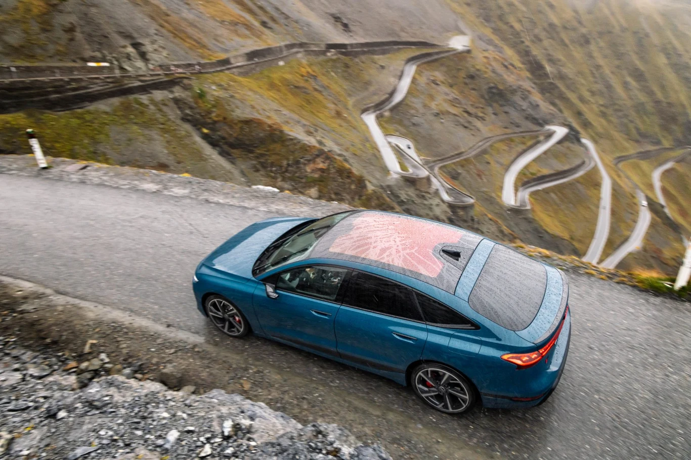 Serpentyny Stelvio widziane z góry. 48 zakrętów, 2758 metrów n.p.m. i elektryczne Audi, które nie zna słowa "stres" Niebieski samochód osobowy poruszający się górską, krętą drogą z licznymi serpentynami, otoczony surowym, skalistym krajobrazem.