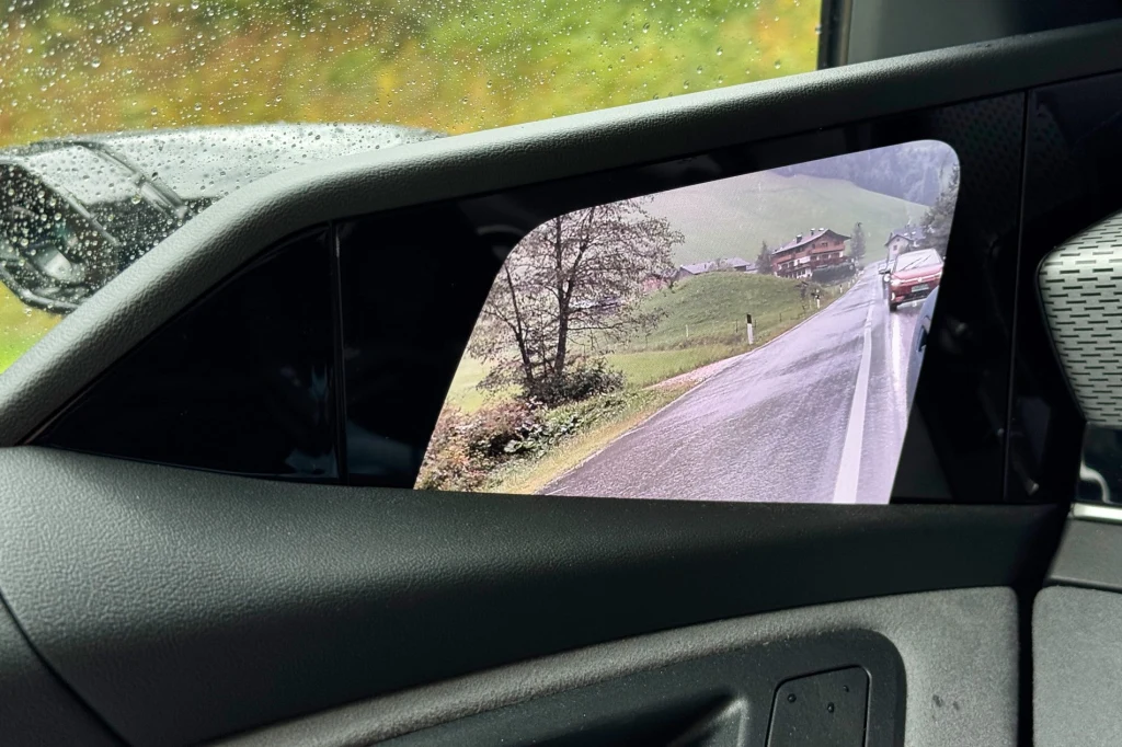 Widok cyfrowego lusterka bocznego w samochodzie, na ekranie widoczna jest droga prowadząca przez tereny wiejskie z domem na wzgórzu, samochodem nadjeżdżającym z naprzeciwka oraz zielenią w tle.