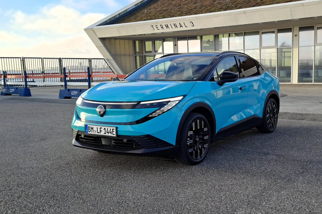 Nissan Leaf mocno się zmienił. Teraz jest crossoverem, ale napęd pozostał bez zmian. Niebieski samochód elektryczny typu SUV zaparkowany przed nowoczesnym budynkiem z dużymi przeszkleniami i napisem Terminal 3, pojazd na niemieckich tablicach rejestracyjnych, asfaltowe podłoże.