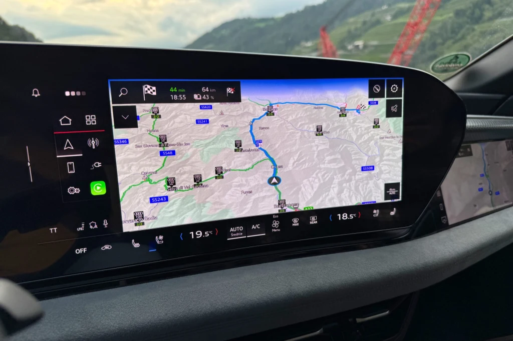 Kolorowy ekran nawigacji samochodowej z wyraźnie zaznaczoną trasą podróży przez górzysty teren, widoczne są szczegółowe informacje o drodze oraz panele kontrolne klimatyzacji i ustawień auta poniżej ekranu.