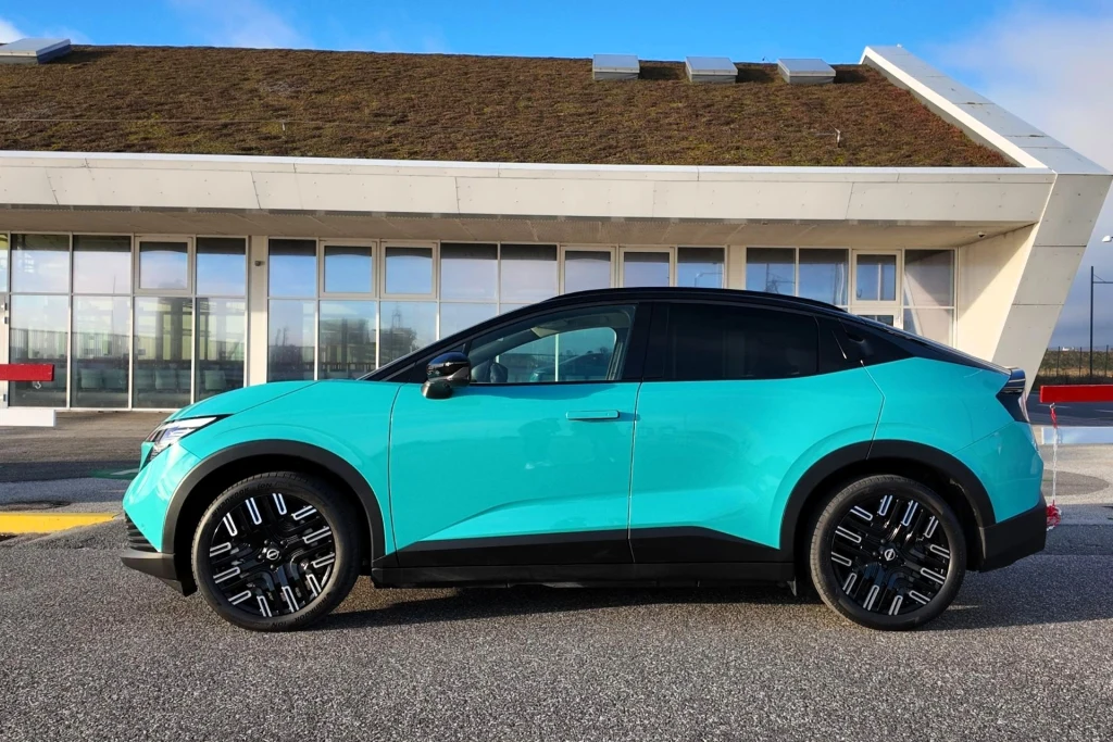 Nowy Nissan Leaf ma 4,35 m długości i 1,55 m wysokości. Rozstaw osi to 2,69 m. Nowoczesny, turkusowy samochód SUV zaparkowany na tle budynku z dużymi oknami i dachem porośniętym roślinnością, wyraźnie zarysowane czarne elementy nadwozia i nowoczesne felgi.