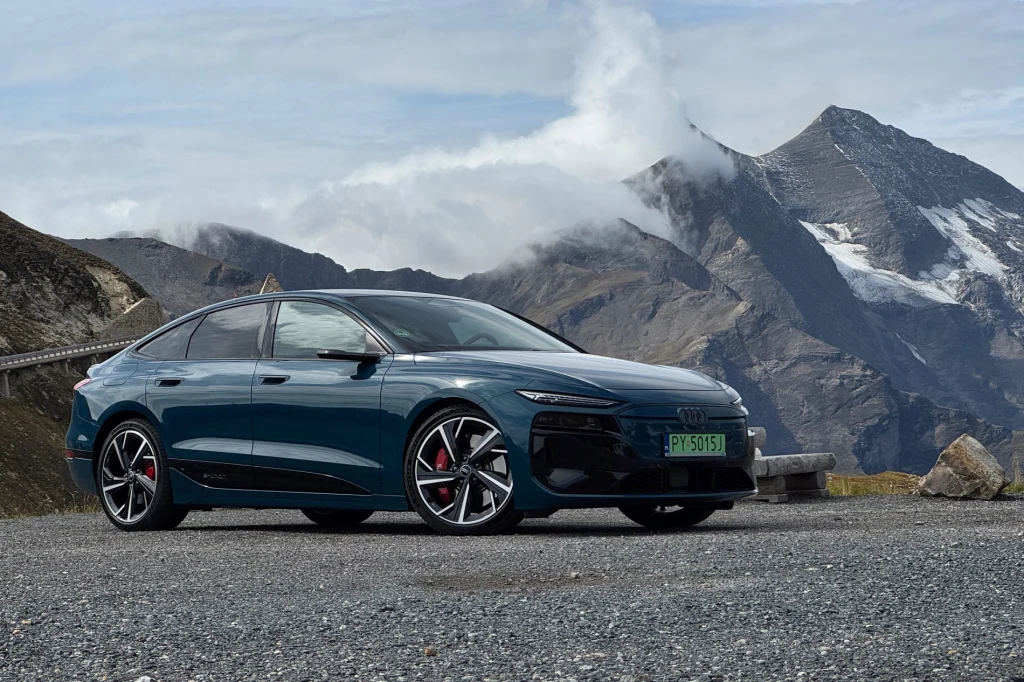 Nowoczesny samochód Audi zaparkowany na tle górskich szczytów i chmur, asfaltowa droga w otoczeniu kamieni i surowego krajobrazu alpejskiego.