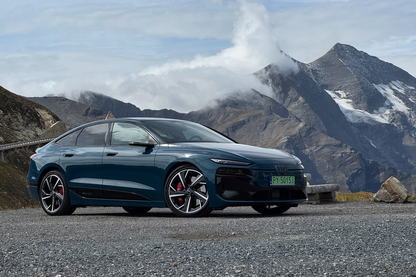 Dojechało z Polski na kołach i na kołach wróci. Zero stresu Nowoczesny samochód Audi zaparkowany na tle górskich szczytów i chmur, asfaltowa droga w otoczeniu kamieni i surowego krajobrazu alpejskiego.