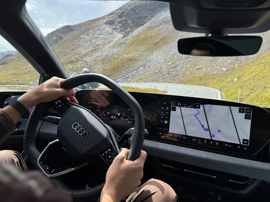 Widok na deskę rozdzielczą samochodu Audi, kierownicę trzymaną przez kierowcę oraz duży wyświetlacz nawigacji pokazujący trasę w górzystym terenie, w tle zakręcająca, asfaltowa droga i górski krajobraz.