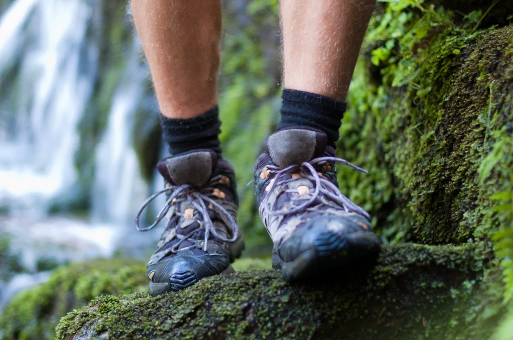 Podeszwy butów trekkingowych zanieczyszczają środowisko podobnie jak opony samochodowe Buty trekkingowe na nogach osoby idącej po porośniętym mchem, wilgotnym kamieniu w otoczeniu przyrody i wodospadu w tle.