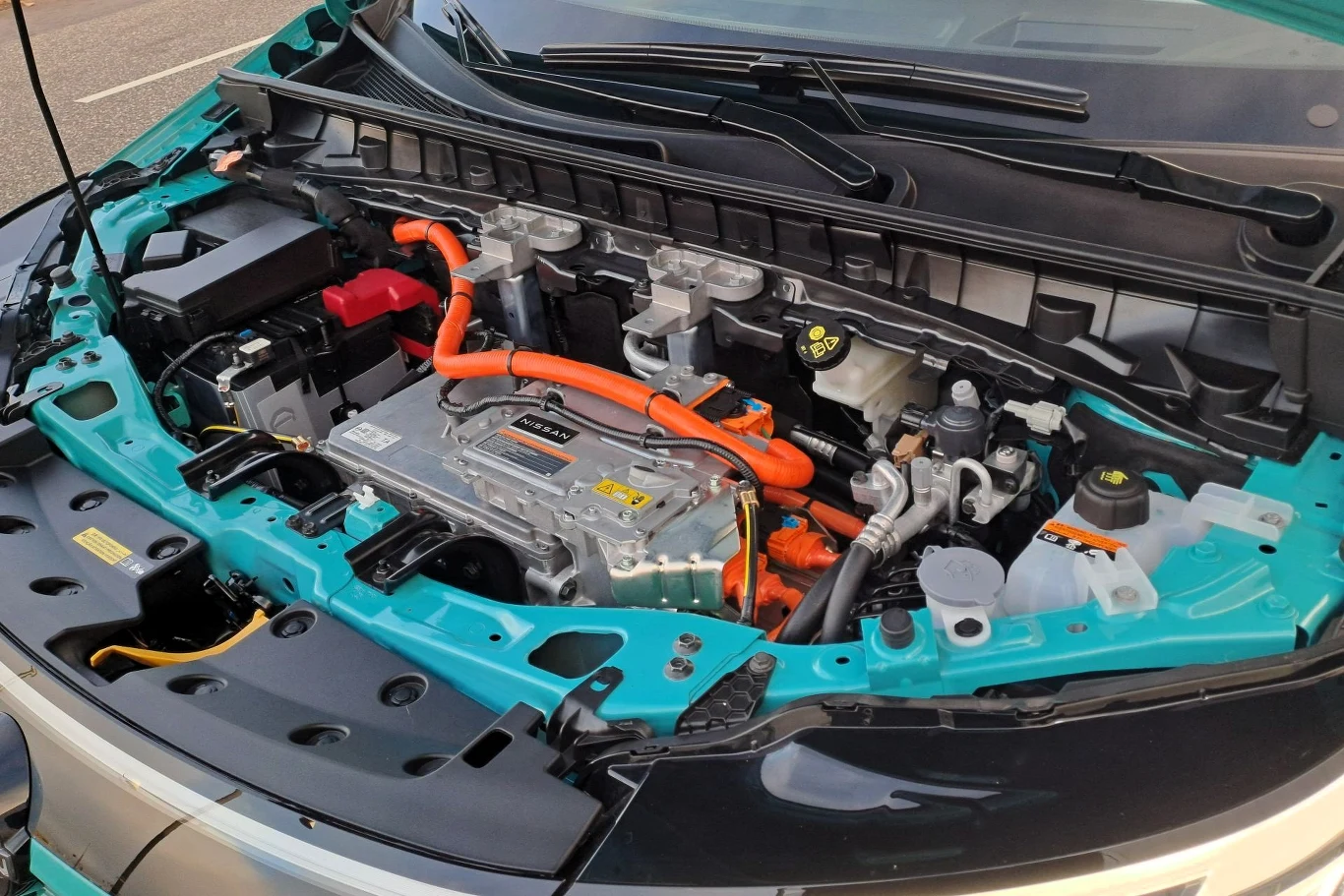 Nissan Leaf dostępny jest w dwóch wariantach mocy: 177 KM i 218 KM. Silnik samochodu elektrycznego z wyeksponowanymi elementami instalacji wysokiego napięcia, widocznymi pomarańczowymi przewodami oraz innymi podzespołami technicznymi pod maską.
