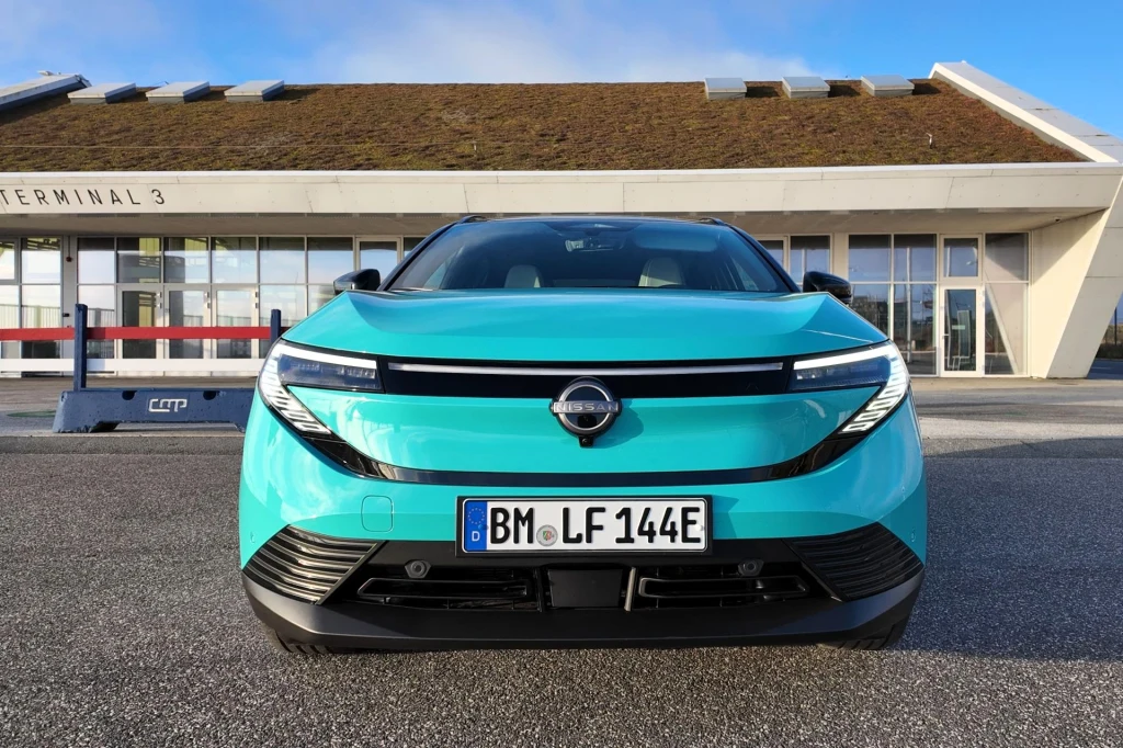 Średnie zużycie Nissana Leafa przy jeździe w okolicach Kopenhagi wyniosło 13,6 kWh/100 km. To mniej niż dane deklarowane przez producenta. Turkusowy samochód marki Opel zaparkowany przodem do aparatu na tle nowoczesnego, przeszklonego budynku z dachem pokrytym roślinnością.