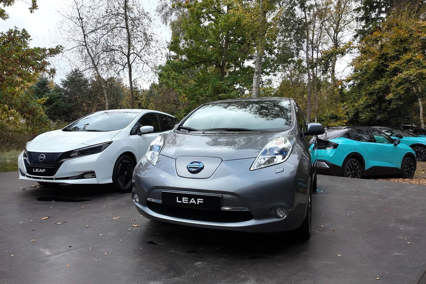 Trzy samochody elektryczne Nissan Leaf zaparkowane na asfaltowej powierzchni w otoczeniu drzew, wśród których są dwa modele w kolorach szarym i białym oraz jeden w kolorze turkusowym, wszystkie oznaczone logo marki.