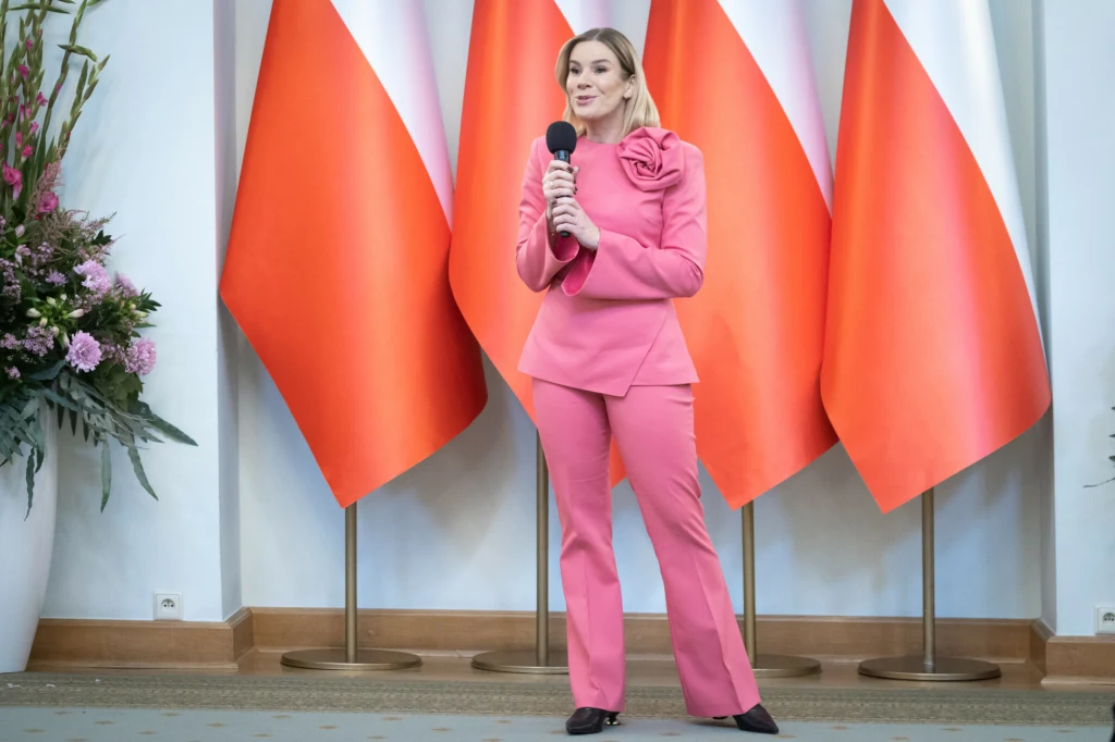 Kobieta w różowym garniturze trzyma mikrofon i stoi przed trzema dużymi polskimi flagami, obok widoczna ozdobna kompozycja kwiatowa w białej donicy.