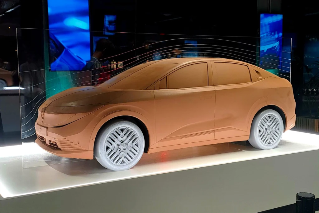 Nissan Leaf trzeciej generacji charakteryzuje się współczynnikiem oporu powietrza Cd na poziomie 0,25. Glina model nowoczesnego samochodu osobowego z białymi kołami ustawiony na podświetlonej ekspozycji, w tle widoczne są monitory i przezroczysta przegroda z liniami symbolizującymi przepływ powietrza wokół pojazdu.