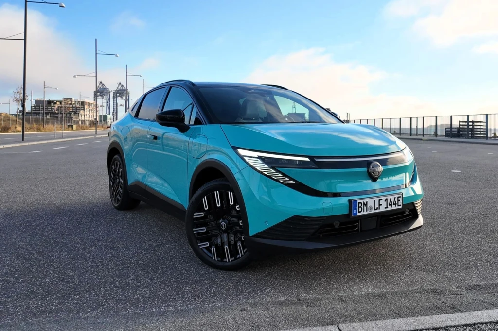 Nissan Leaf dostępny jest z dwoma wariantami akumulatora: 52 kWh i 75 kWh. Ten drugi zapewnia zasięg na jednym ładowaniu do 620 km. Nowoczesny, turkusowy samochód elektryczny zaparkowany na pustym parkingu, na tle miejskiej architektury oraz jasnego, częściowo zachmurzonego nieba.
