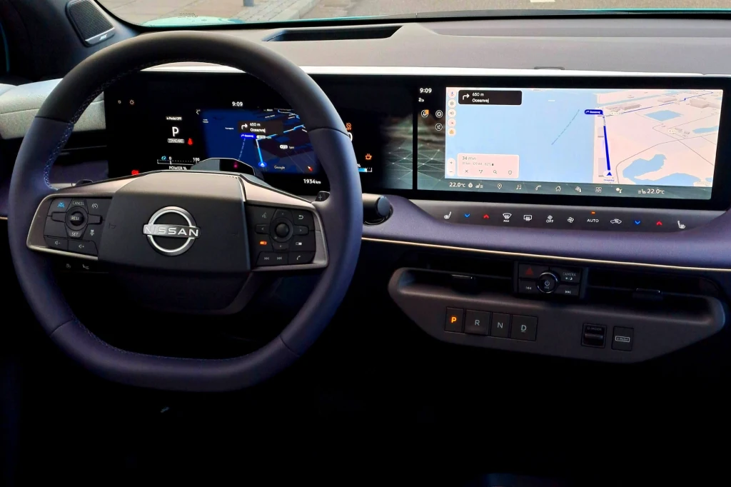 Nissan Leaf wyposażony jest w dwa optycznie połączone ze sobą ekrany o przekątnej 14,3 cala. Kokpit nowoczesnego samochodu marki Nissan z widocznym logo na kierownicy, dużym cyfrowym wyświetlaczem za kierownicą oraz szerokim ekranem multimedialnym z nawigacją i cyfrową deską rozdzielczą.