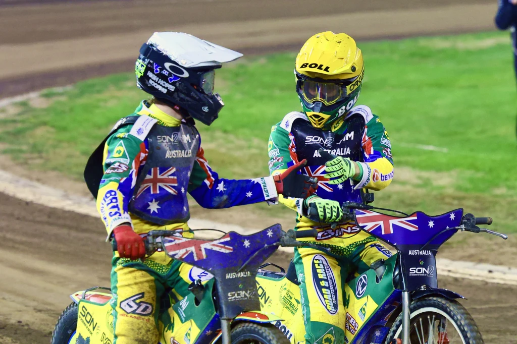 Tate Zische i Mitchell McDiarmid w finale SON2. Dwóch australijskich żużlowców ubranych w kombinezony w barwach narodowych ściska sobie ręce, stojąc na motocyklach na torze żużlowym, na tle fragmentu zielonej murawy.