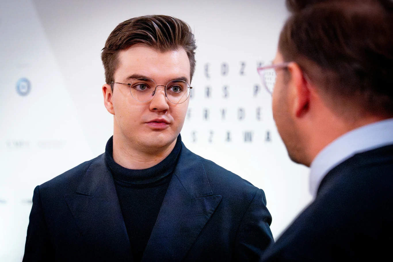 Młody mężczyzna w okularach i ciemnej marynarce rozmawia z drugim mężczyzną, który widoczny jest częściowo z profilu, w tle rozmazane litery testu okulistycznego.