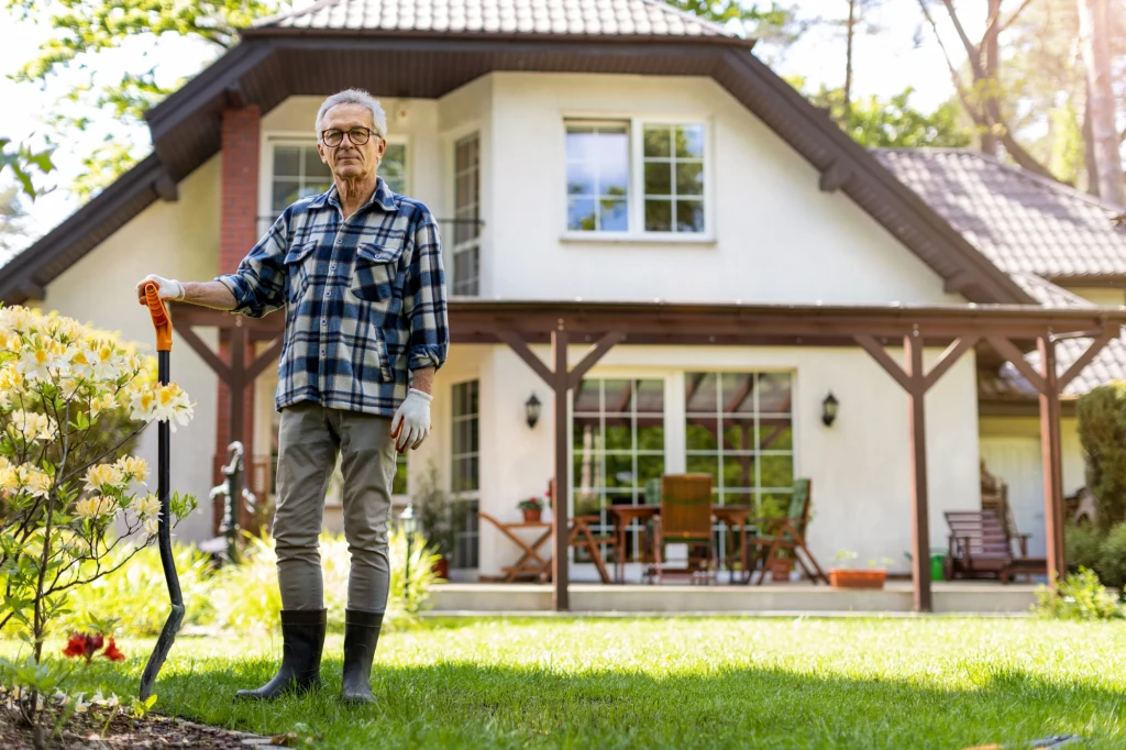 Kto dziedziczy spadek po bezdzietnym? Aby odpowiedzieć, trzeba sięgnąć do prawa spadkowego. Starszy mężczyzna w okularach i kaloszach stojący na zielonym trawniku przed domem jednorodzinnym, trzymający w ręku łopatę do ogrodu, dookoła zadbane rabaty i ogród.