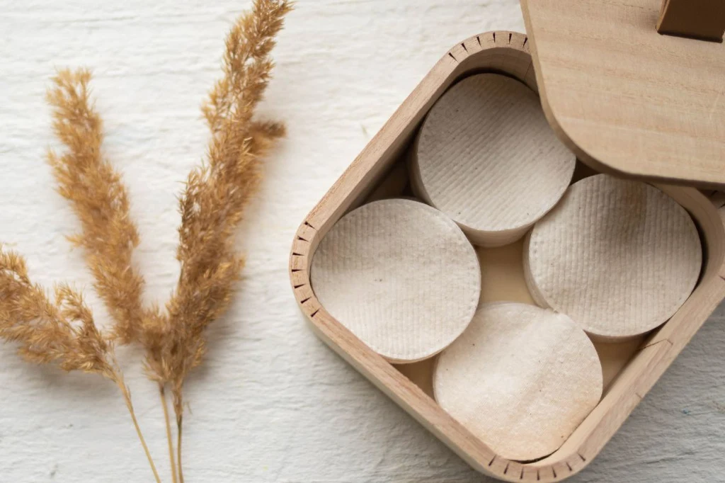 Wystarczą płatki kosmetyczne, trochę pudru i zamykany pojemnik Cztery okrągłe drewniane krążki w otwartym pudełku obok suszonych traw na jasnym tle.