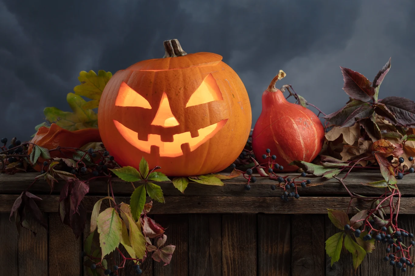 Halloweenową dynię możemy ozdobić techniką etchingu. Wydrążona dynia z wyciętym uśmiechniętym wzorem i świecącym środkiem ustawiona na drewnianym ogrodzeniu, obok mniejsza dynia i jesienne liście, w tle ciemne, burzowe niebo.