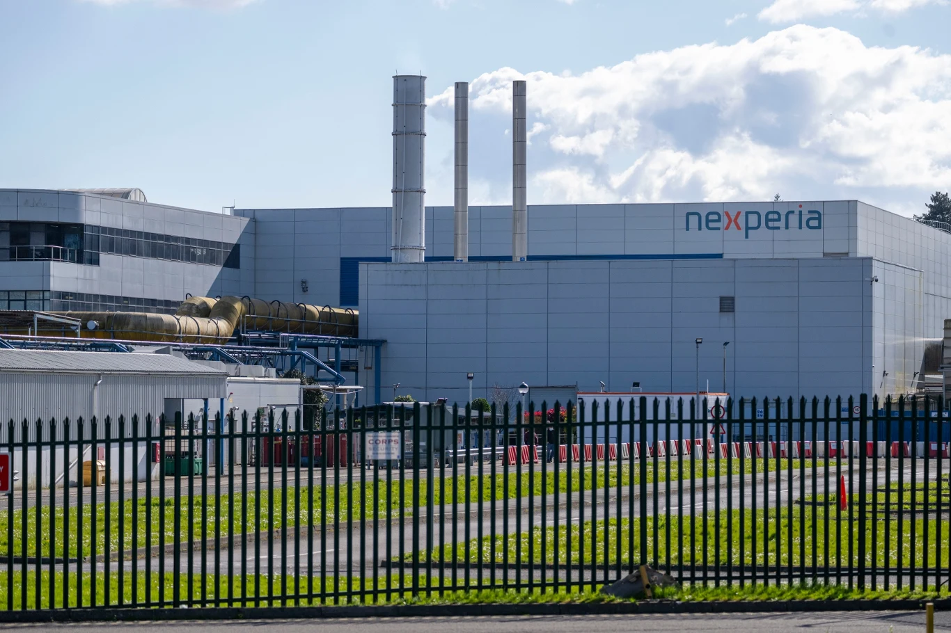 Nowoczesny budynek przemysłowy z wyraźnym logo firmy nexperia, otoczony metalowym ogrodzeniem, w tle widoczne trzy wysokie kominy oraz nieco zachmurzone niebo.