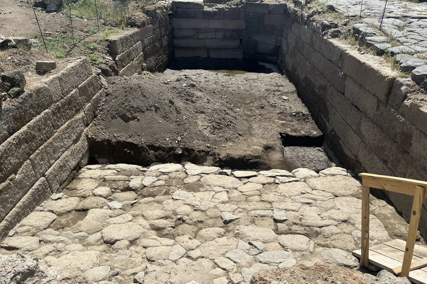 W sercu starożytnego rzymskiego miasta Gabii, położonego zaledwie 18 kilometrów na wschód od Rzymu, archeolodzy odkryli pozostałości ogromnego, wyłożonego kamieniem basenu, częściowo wykutego bezpośrednio w podłożu skalnym. Kamienna wykopaliska archeologiczne z odsłoniętymi fragmentami murów oraz brukowanej drogi, otoczone wysokimi, prostokątnymi blokami kamiennymi, z obecnymi śladami ziemi i drewnianą balustradą w prawym dolnym rogu.