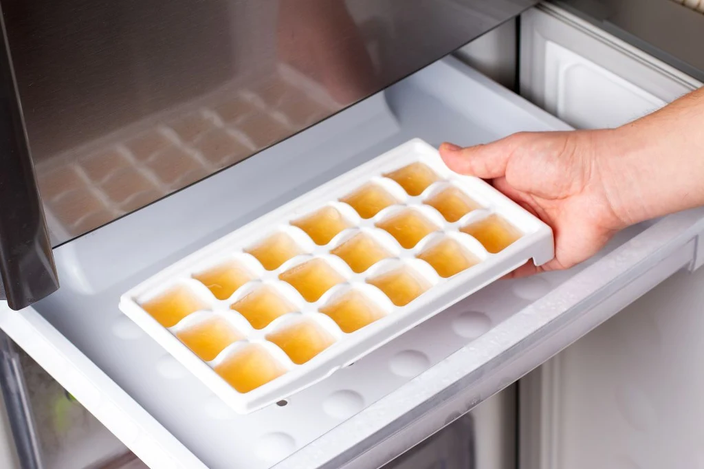 Dzięki mrożeniu, zdrowe kostki na przeziębienie możemy mieć pod ręką cały sezon Tacka z zamrożonymi kostkami o nietypowym, lekko brązowym kolorze umieszczona w zamrażarce, trzymana przez dłoń, widoczny fragment wnętrza urządzenia chłodniczego.