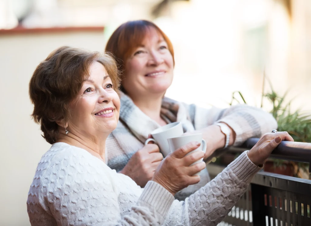 Dwie starsze kobiety uśmiechają się i trzymają kubki z napojem na balkonie, ubrane w jasne swetry, w tle widoczna roślinność.
