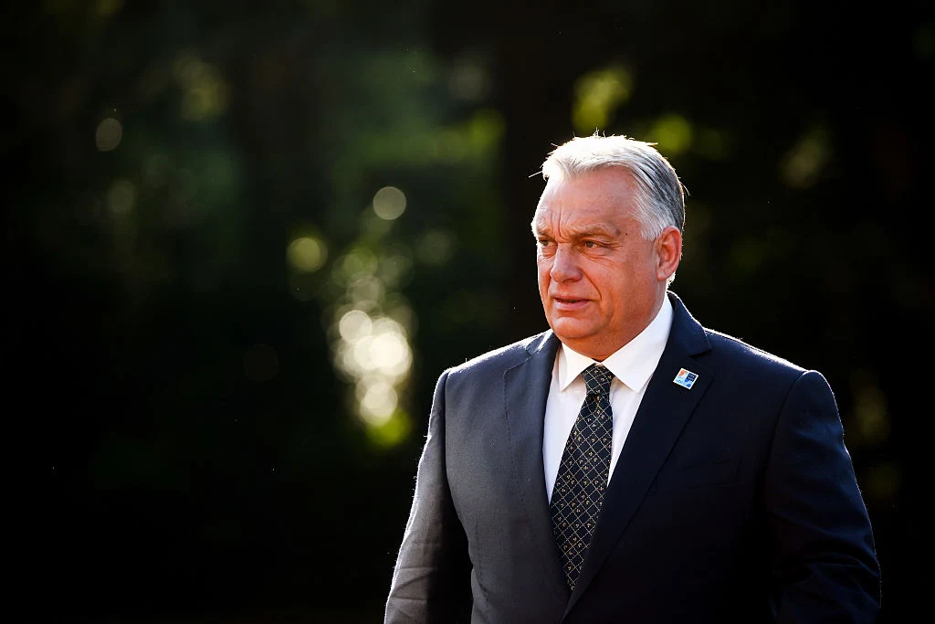 Premier Węgier Viktor Orban Mężczyzna w średnim wieku ubrany w ciemny garnitur z krawatem, stojący na tle rozmytego, zielonego krajobrazu, światło słoneczne pada na jego twarz, co podkreśla powagę wyrazu.