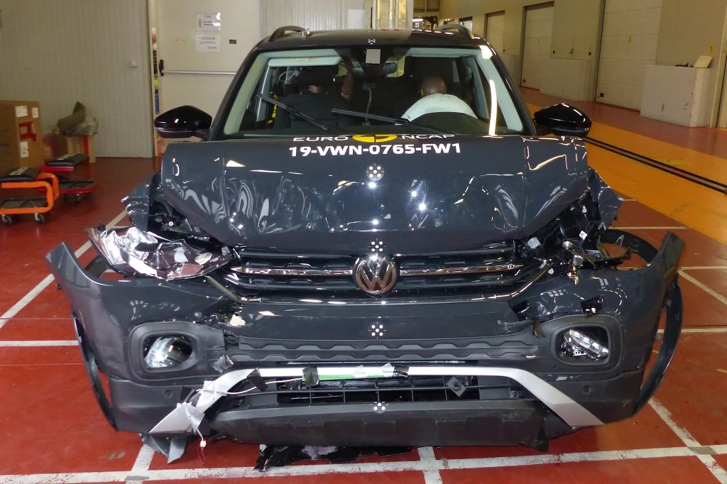 Uszkodzony przód samochodu Volkswagen po teście zderzeniowym, widoczne zgniecenia maski, zderzaka oraz reflektorów, pojazd ustawiony na oznaczonej strefie w hali testowej, na szybie numer testowy Euro NCAP.