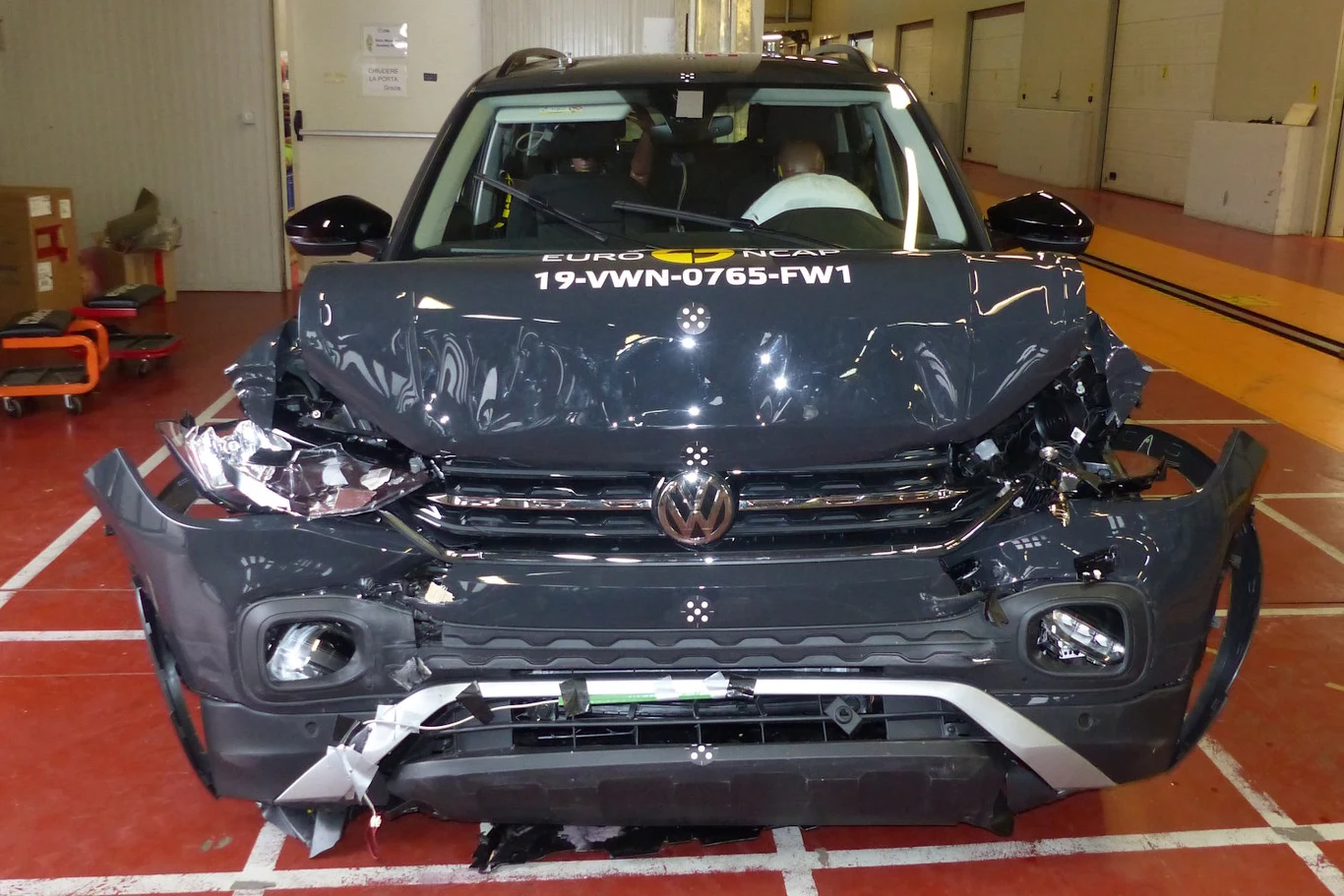 Uszkodzony przód samochodu Volkswagen po teście zderzeniowym, widoczne zgniecenia maski, zderzaka oraz reflektorów, pojazd ustawiony na oznaczonej strefie w hali testowej, na szybie numer testowy Euro NCAP.