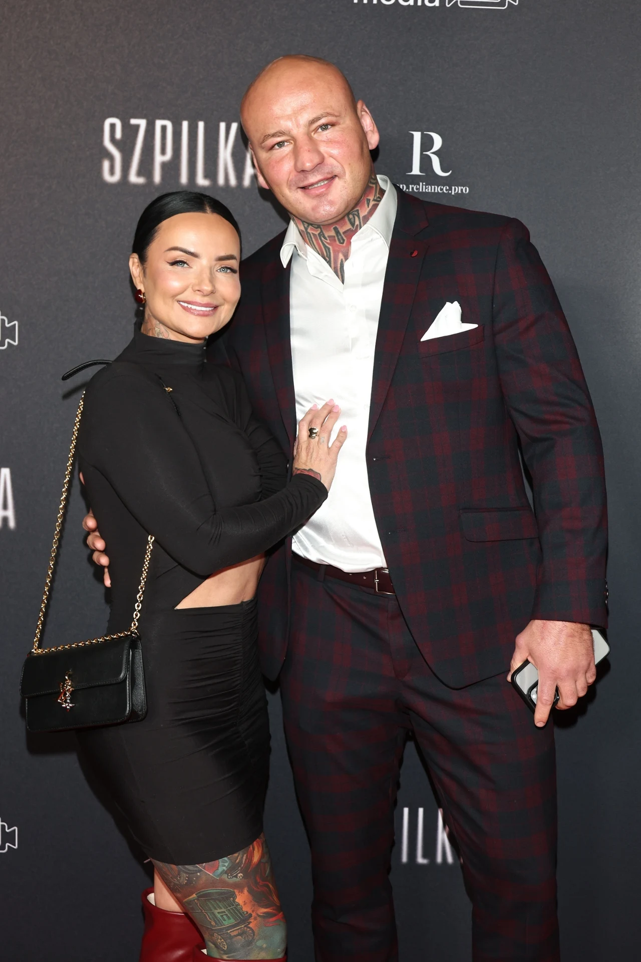 Kobieta i mężczyzna stoją razem na tle ściany fotograficznej z nazwą wydarzenia, mężczyzna w eleganckim garniturze, a kobieta w czarnej sukience z torebką na ramieniu, oboje uśmiechnięci.