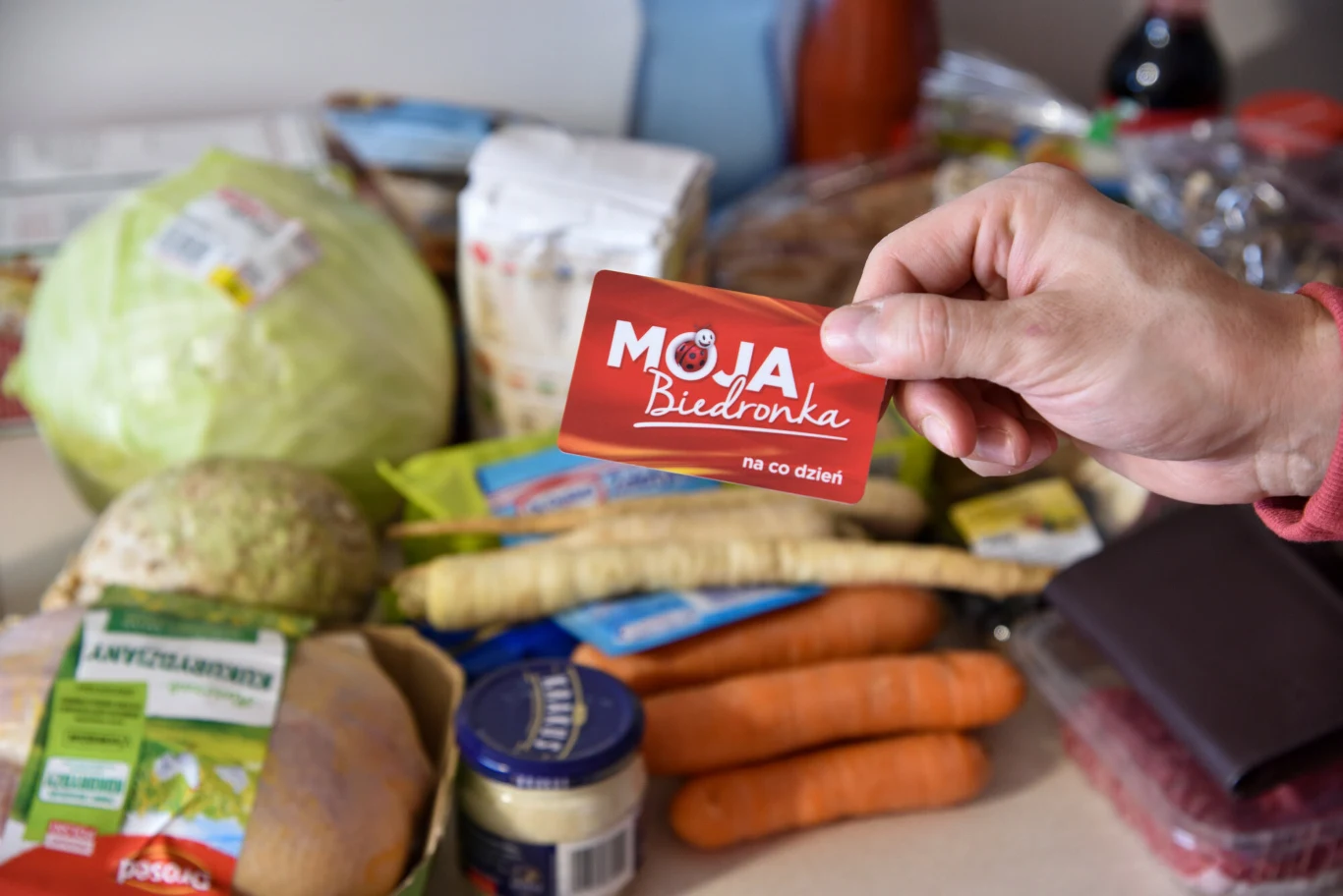 Usuwasz konto Biedronka bez zastanowienia. Możesz stracić dostęp do promocji (zdj. ilustracyjne) Ręka trzymająca kartę lojalnościową Moja Biedronka na tle różnych produktów spożywczych, takich jak marchewki, kapusta, chrzan, chleb, ogórek konserwowy, ser oraz inne artykuły spożywcze.