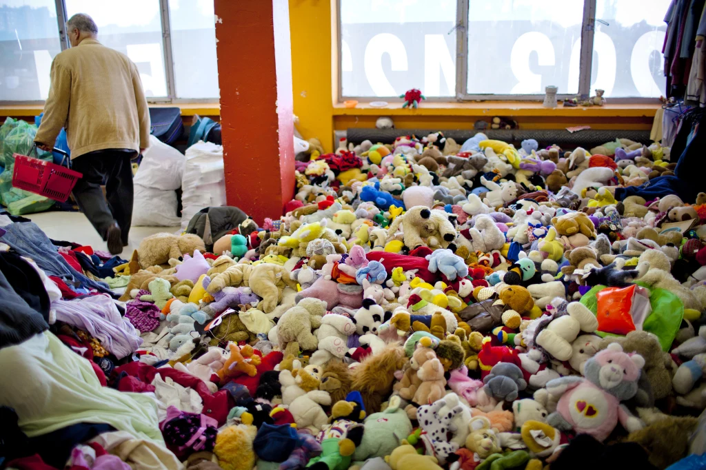 W skepach z tanią odzieżą można było kupić nie tylko ubrania Ogromna liczba pluszowych zabawek rozrzuconych na podłodze dużego jasnego pomieszczenia, z lewej strony starszy mężczyzna odchodzi, trzymając czerwoną torbę. W tle widać stosy ubrań oraz duże okna.