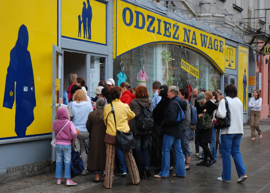 Do lumpeksów wciąż warto zaglądać Grupa ludzi czekająca przed wejściem do sklepu z odzieżą na wagę, tło stanowi żółto-niebieska witryna sklepu, w oknie dostrzegalne są manekiny ubrane w kolorowe ubrania, chodnik jest mokry po deszczu.