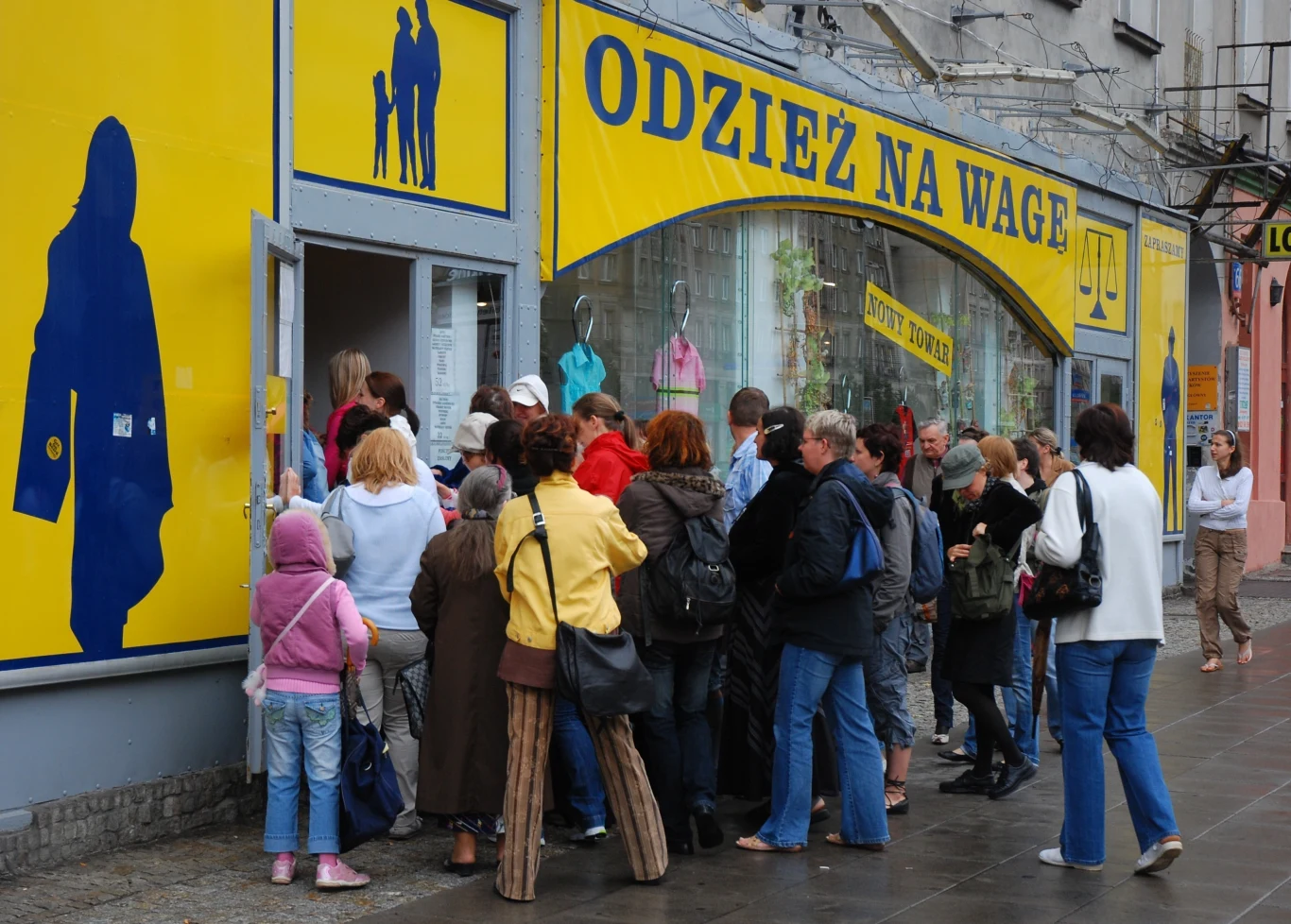 Do lumpeksów wciąż warto zaglądać Grupa ludzi czekająca przed wejściem do sklepu z odzieżą na wagę, tło stanowi żółto-niebieska witryna sklepu, w oknie dostrzegalne są manekiny ubrane w kolorowe ubrania, chodnik jest mokry po deszczu.