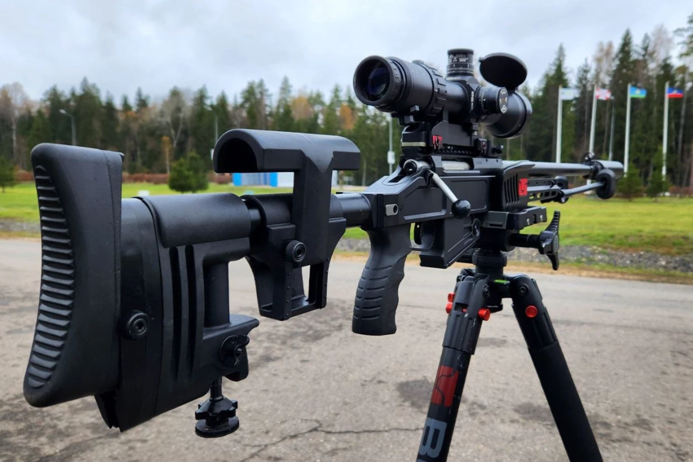 Rosja wysyła nowe karabiny snajperskie żołnierzom na froncie w Ukrainie Karabin snajperski wyposażony w lunetę celowniczą zamocowany na statywie, w tle widoczny pas zieleni, drzewa oraz kilka masztów z flagami.