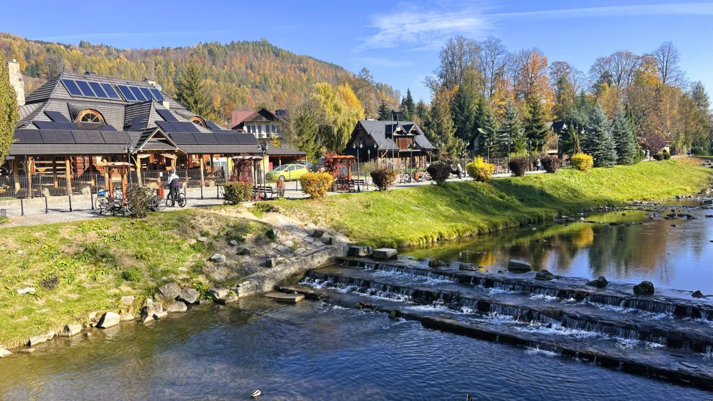 Przejażdżka promenadą nad Grajcarkiem jest prawdziwą przyjemnością Rzeka z kaskadami wodnymi płynąca przez wieś, po lewej stronie nowoczesne drewniane budynki z panelami słonecznymi i rowerzyści, w tle jesienny las i góry.