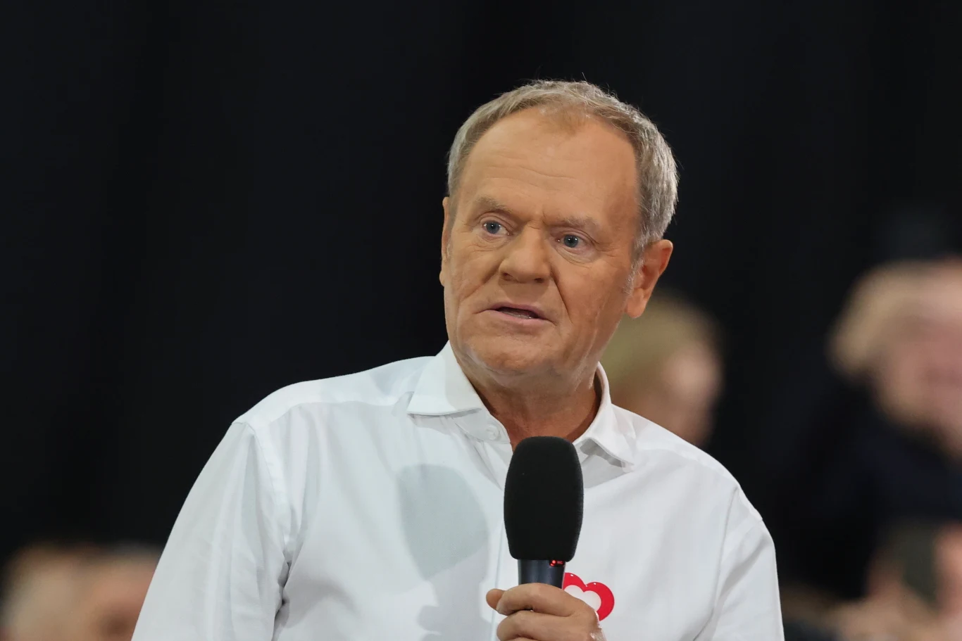 Mężczyzna trzymający mikrofon, ubrany w białą koszulę z nadrukowanym wzorem serca, stoi na tle rozmazanej publiczności.