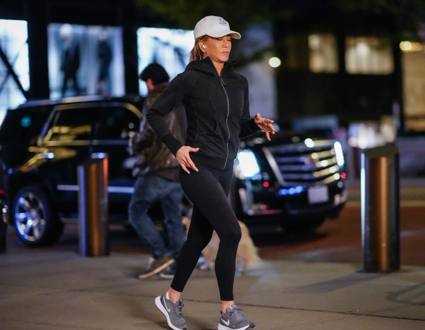 Jennifer Aniston jest niezwykle aktywna i nie ma zamiaru przestawać dbać o swoje zdrowie w ten sposób Kobieta w sportowym stroju, czapce z daszkiem i butach do biegania spaceruje lub biega po chodniku miejskim, w tle widać zaparkowany czarny samochód typu SUV, a także innych przechodniów.
