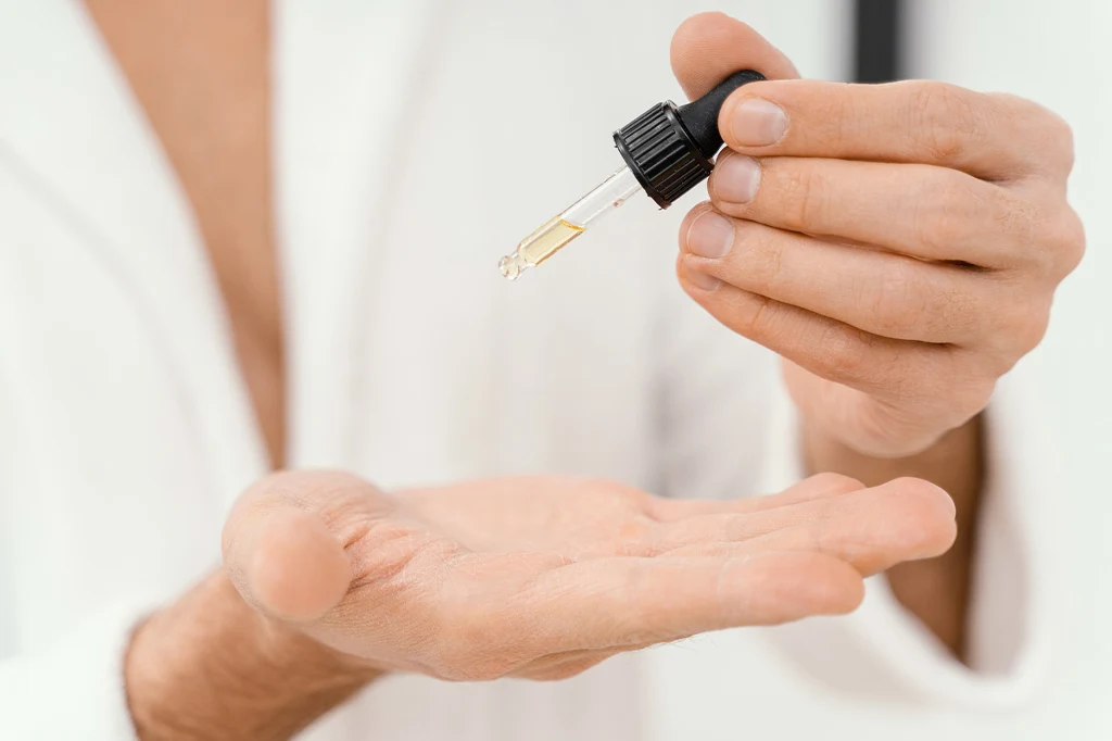 Ręce osoby aplikującej krople lub olejek z pipety na otwartą dłoń, ujęcie skupione na dłoniach i akcesorium kosmetycznym. Ręce osoby aplikującej krople lub olejek z pipety na otwartą dłoń, ujęcie skupione na dłoniach i akcesorium kosmetycznym.