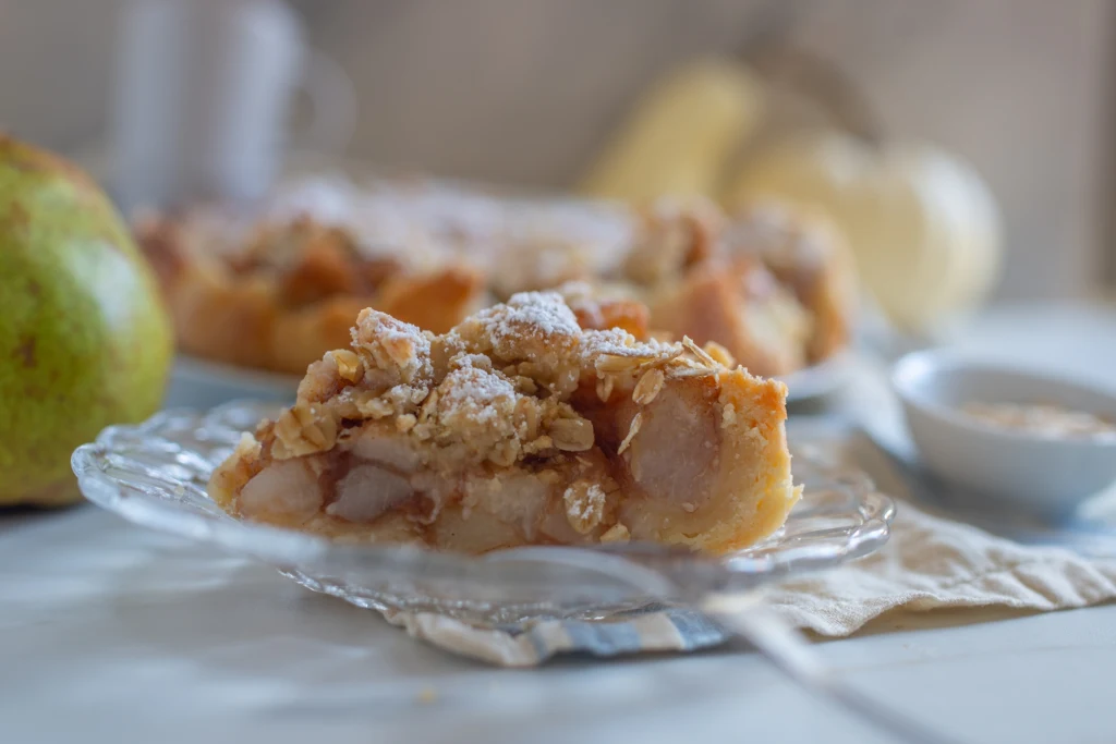 Kawałek szarlotki z kruszonką i cukrem pudrem, podany na przezroczystym szklanym talerzyku, w tle widoczny kawałek gruszki i rozmyte elementy zastawy stołowej.