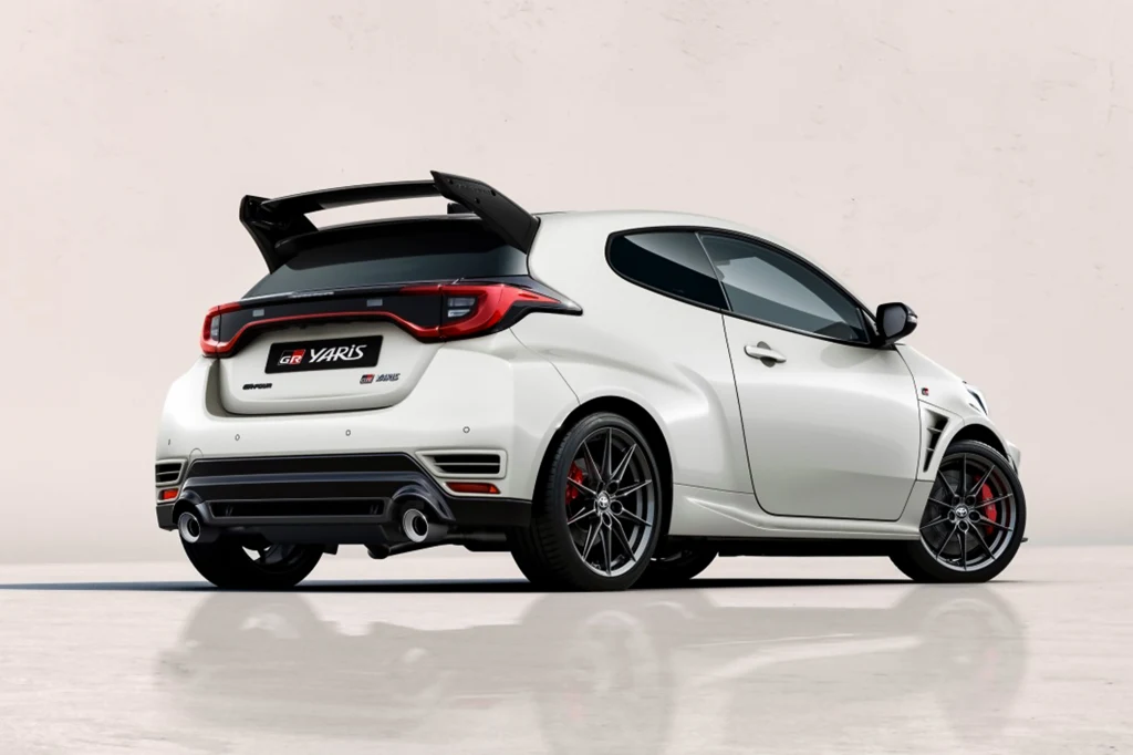 Toyota GR Yaris w wersji Aero Performance Biały sportowy samochód Toyota GR Yaris z dużym tylnym spojlerem, czerwonymi detalami na zderzaku i felgami w ciemnym kolorze, stojący na gładkiej powierzchni na jednolitym tle.