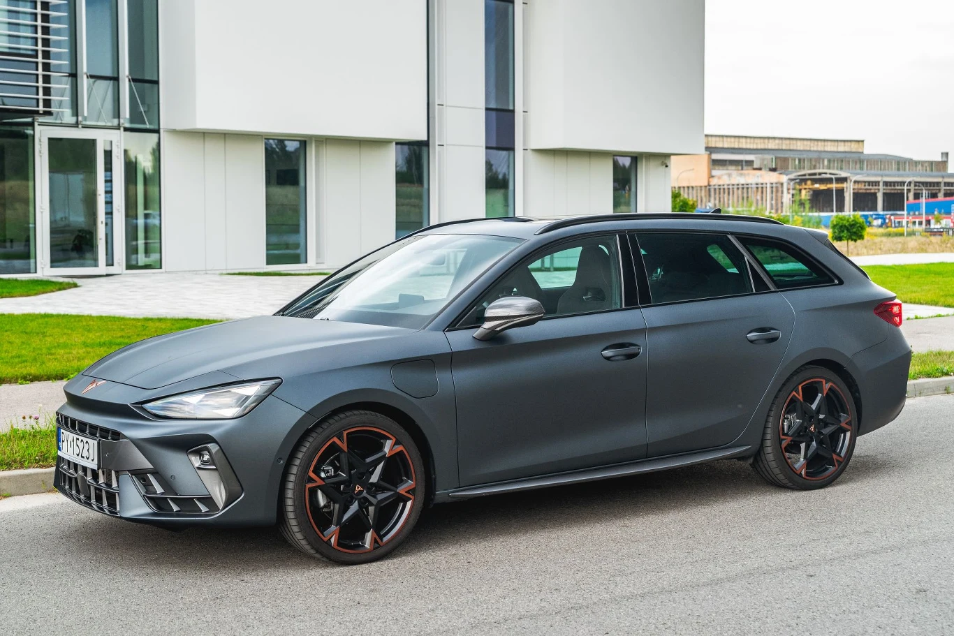 Cupra Leon z nową hybrydą plug-in jest wolniejsza, ale oszczędniejsza. Ciemnoszary samochód kombi z przyciemnianymi szybami zaparkowany przy chodniku na tle nowoczesnego budynku z dużymi oknami, czarne felgi z pomarańczowymi akcentami podkreślają sportowy charakter pojazdu.