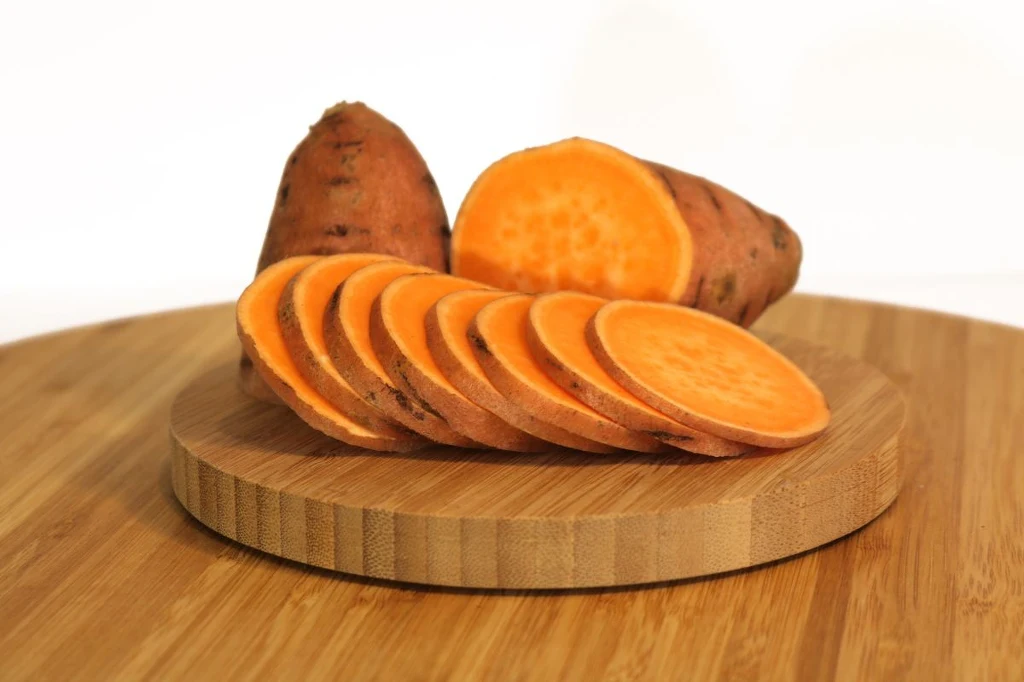 Słodkie ziemniaki, czyli bataty doskonale zastąpią klasyczne ziemniaki Pokrojony batat ułożony na okrągłej, drewnianej desce do krojenia, w tle widoczna większa część niepokrojonego batata.