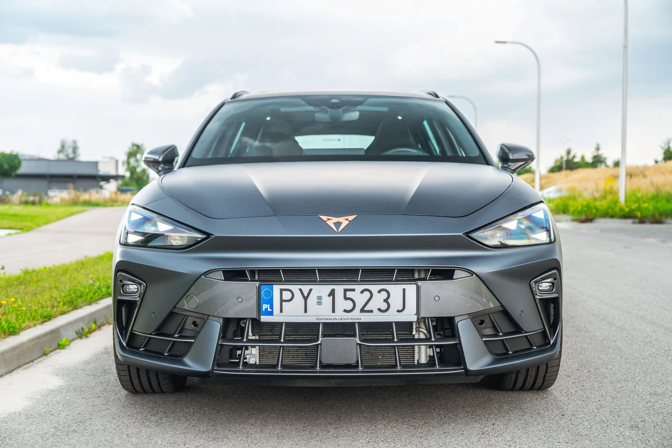 Po modernizacji Cupra Leon otrzymała nowy bardziej wyrazisty przód. Odróżnia się teraz od modelu Seata. Czarny samochód Cupra o nowoczesnym, sportowym designie widoczny od przodu, z polskimi tablicami rejestracyjnymi, stoi na asfaltowej drodze przy krawężniku na tle osiedlowej zabudowy i zieleni.