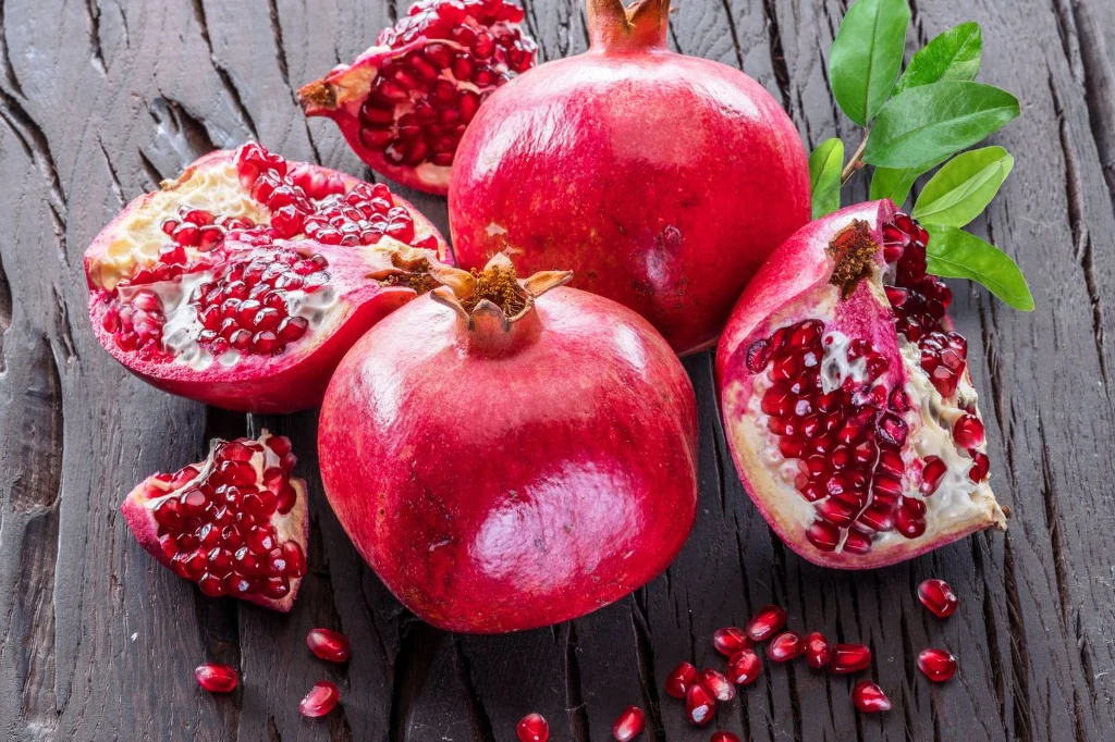 Sok z granatów wielu uważa za najsmaczniejszy ze wszystkich Trzy granaty, dwa w całości i jeden rozkrojony, leżą na drewnianym stole obok kilku zielonych liści, widoczne są błyszczące czerwone pestki zarówno wewnątrz owocu, jak i rozsypane na stole.