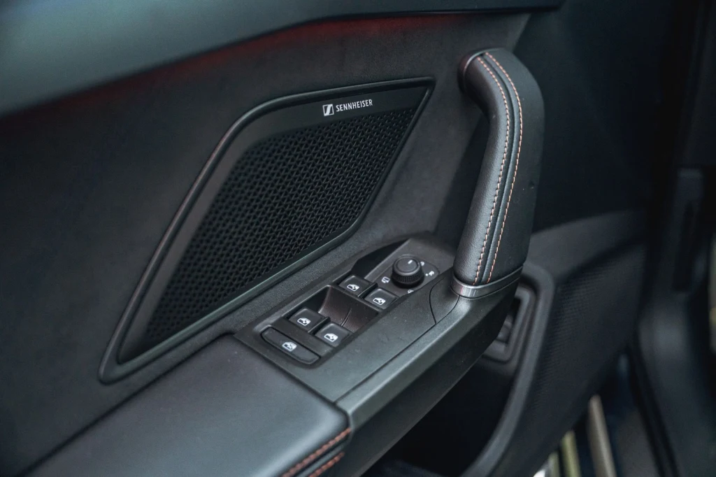 O nagłośnienie może dbać system audio opracowany we współpracy z firmą Sennheiser. panel drzwiowy samochodu z widocznymi przyciskami do sterowania szybami i lusterkami bocznymi, podłokietnikiem z czerwonymi przeszyciami oraz głośnikiem marki Bang & Olufsen