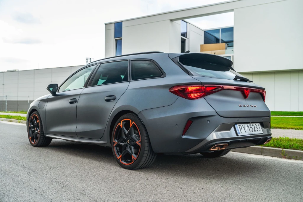 Z uwagi na obecność większego akumulatora Cupra Leon stała się wolniejsza, nawet w mocniejszym, 272-konnym wariancie. Szare, sportowe kombi marki Cupra zaparkowane przy ulicy, wyróżnia się nowoczesnym designem, czerwonymi elementami felg oraz dynamiczną sylwetką na tle nowoczesnego budynku.
